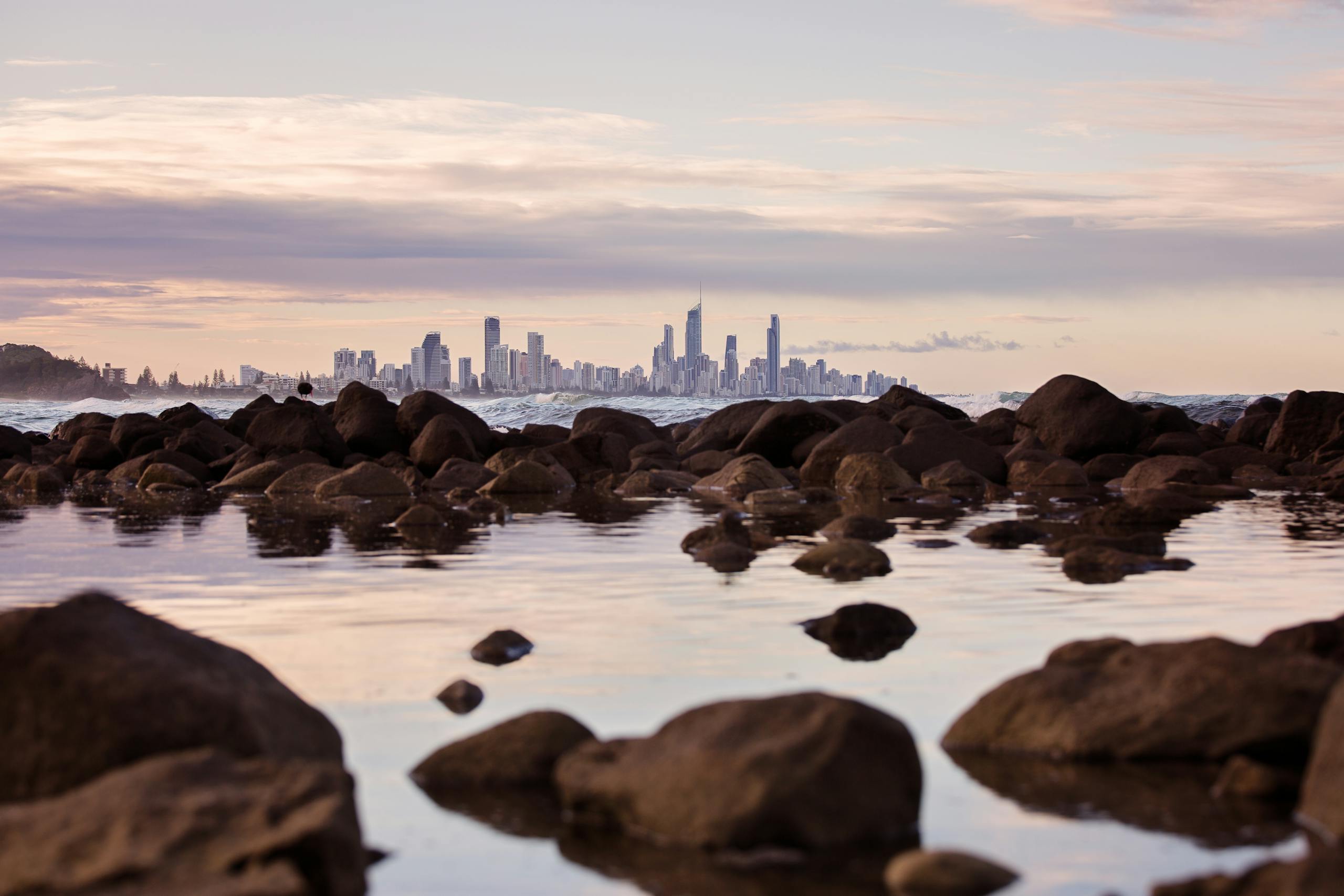 QLCredit Dramatic view of Gold Coast skyline from a rocky shore during a tranquil sunset.