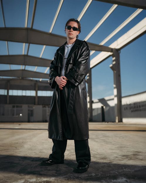 Dis Fashion Man wearing sunglasses and a leather coat standing confidently under skeletal building frame.
