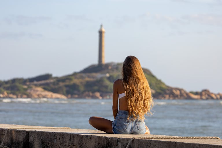 Kulfiy Ke Awaz: Yaadon, Young woman sitting by the sea looking at a distant lighthouse on a sunny day.