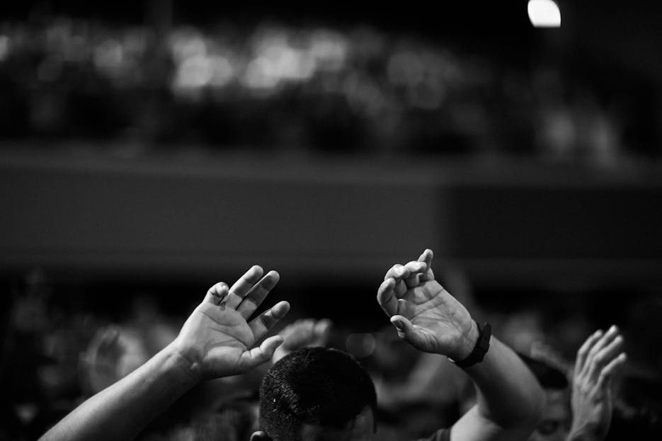 ksözcü A black and white photo of hands raised in a crowd during a concert or religious gathering indoors.
