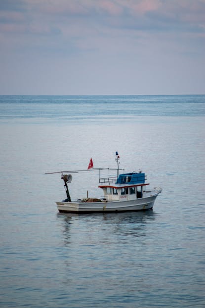 Calmered A lone fishing boat floats peacefully on a calm blue sea under a pastel sky.