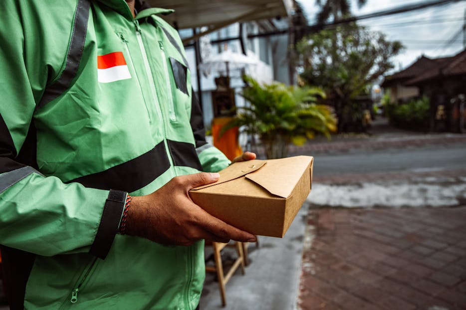 Magento Service Gonzay A person in a green jacket delivering a package outdoors. Urban setting, daytime.