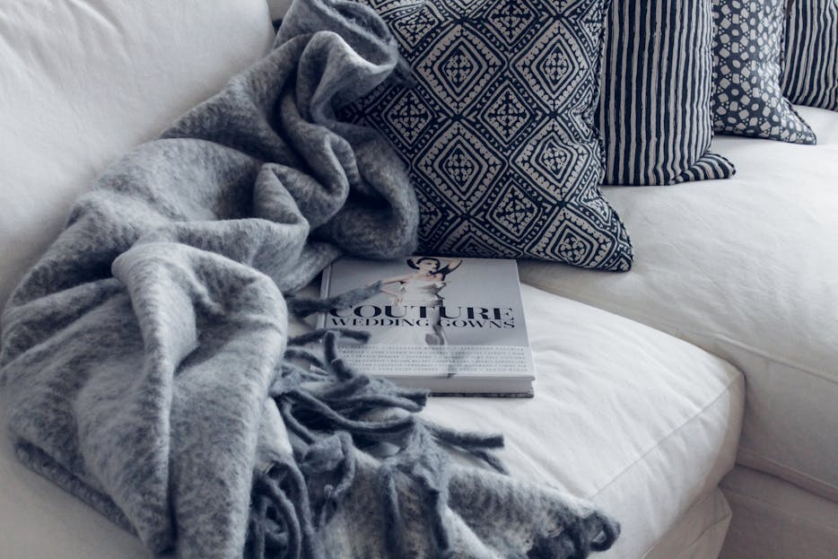 Sofoximmo A stylish living room featuring a white sofa with patterned pillows, a blanket, and a fashion book.