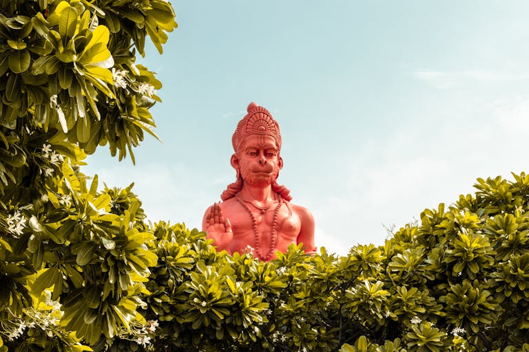 Shri Hanuman Dham A vibrant red statue of Hanuman surrounded by green foliage under a clear sky.