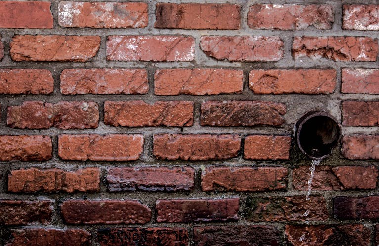 Trencher for Drainage Close-up of a red brick wall with water flowing from a drain, showcasing texture and patterns.