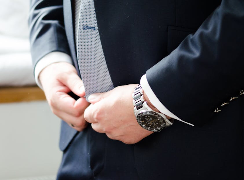 Blazertje Close-up of groom adjusting tie, wearing a formal suit with watch, indoors.