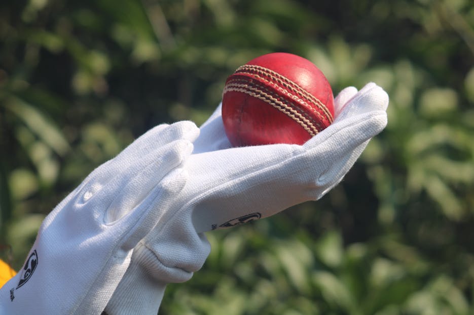 Wheon Cricket 07 Detailed photo of a red cricket ball held by gloved hands outdoors.