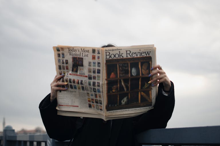 Dainik Shiksha Update News Today: Individual holding a book review newspaper with colorful nails against a cloudy sky backdrop.