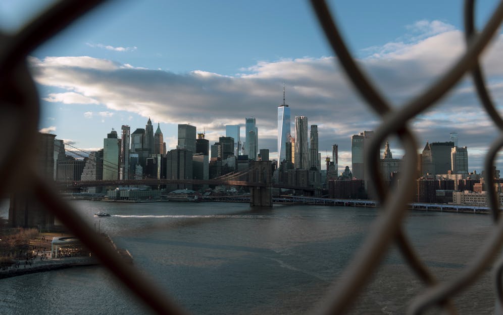 Panorama of World Alaikas Stunning view of New York City skyline through a fence, showcasing skyscrapers and Brooklyn Bridge.