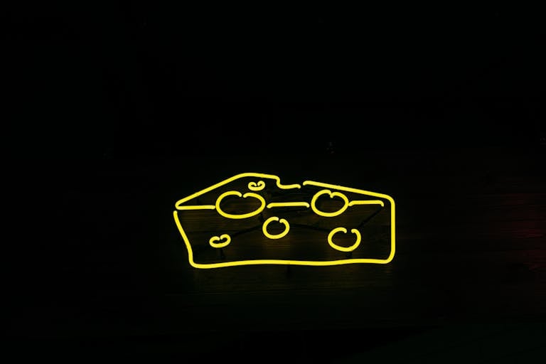 Kulfiy com A bright yellow neon sign shaped like cheese against a black background, creating a modern and eye-catching design.