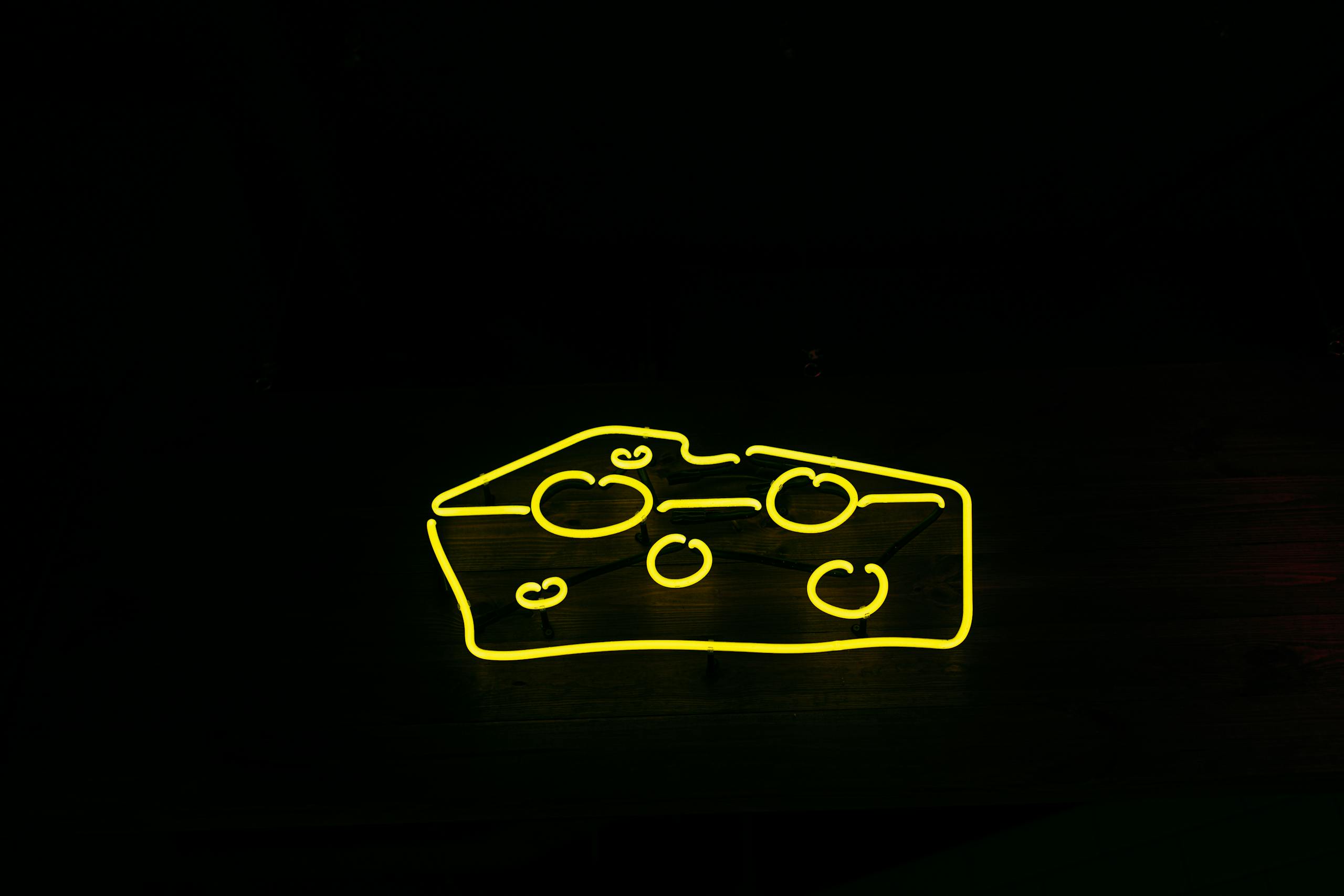 Kulfiy com A bright yellow neon sign shaped like cheese against a black background, creating a modern and eye-catching design.