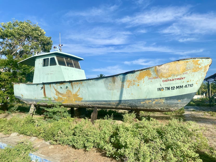 Pak vs Ind A weathered, old fishing boat resting on land surrounded by greenery, under a bright blue sky.