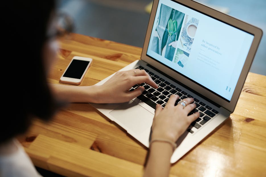 ibomma1.co Person typing on a laptop at a wooden table with a smartphone nearby, illustrating remote work.