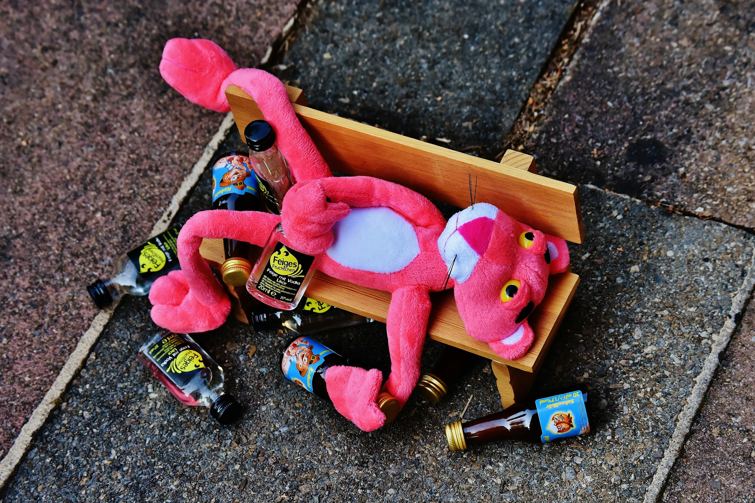 Voozon A pink plush toy sprawled on a bench with mini liquor bottles around it, creating a humorous scene.