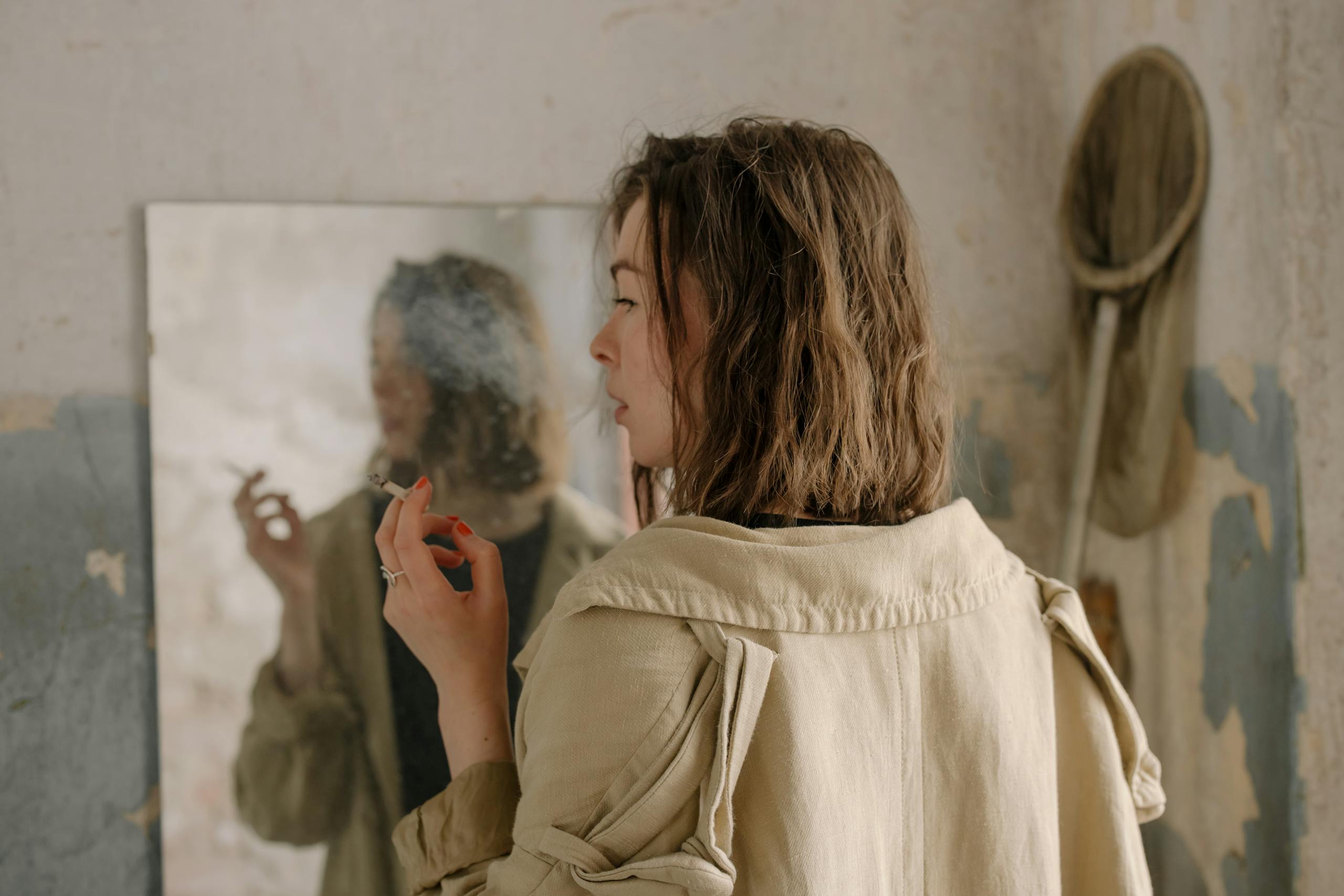 Carmela Clouth Carmela Clouth A woman in a trench coat smoking, reflected in a mirror against a shabby, textured wall.