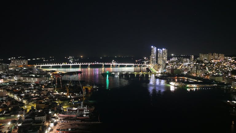 The Vzusa Stunning night view of Busan's illuminated skyline and bridge reflecting on the water.