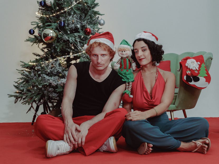 Twastia.com.au A cheerful couple in Santa hats sitting by a Christmas tree with holiday decorations.