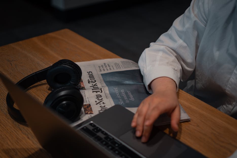 IntelligentNews.co.uk A person works on a laptop with headphones and a newspaper on a wooden table.