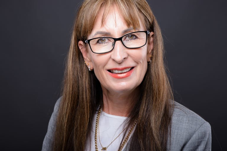 Jan Ashley A senior woman smiling in eyeglasses and a gray coat, with a confident expression.