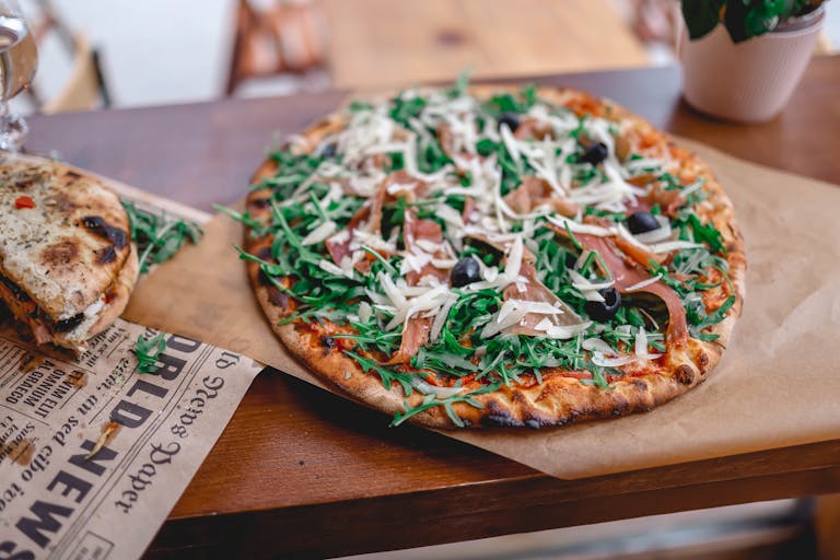 Pizzerija Plus A tasty prosciutto and arugula pizza served on a wooden table, perfect for food photography enthusiasts.