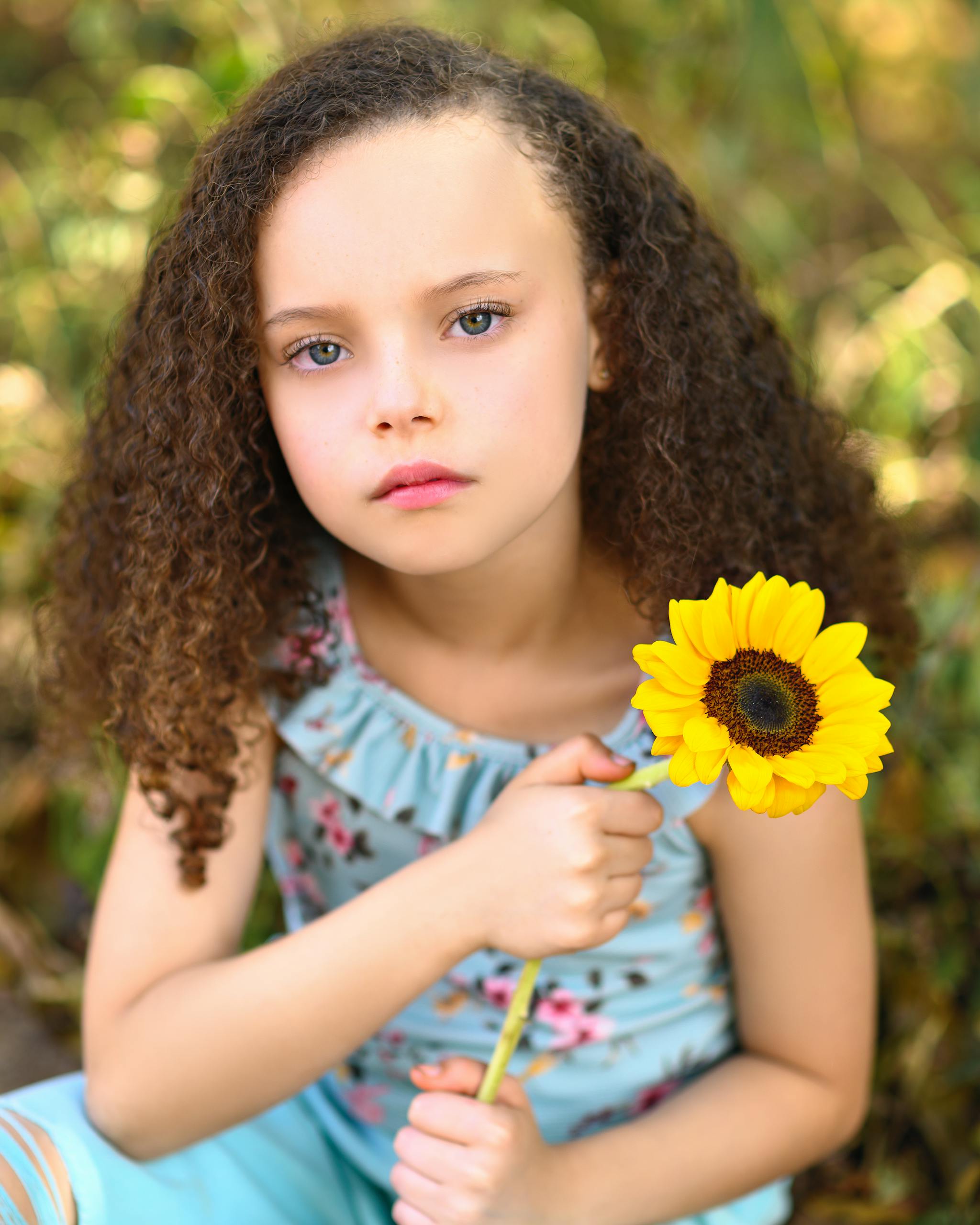 Finley Elaine Griffin A young girl sitting outdoors holding a sunflower, evoking simplicity and innocence.