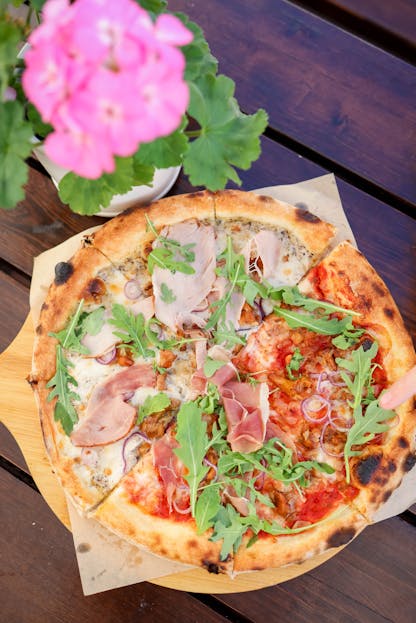 Pizzerija Plus Delicious Italian pizza topped with prosciutto and fresh arugula on a wooden board.