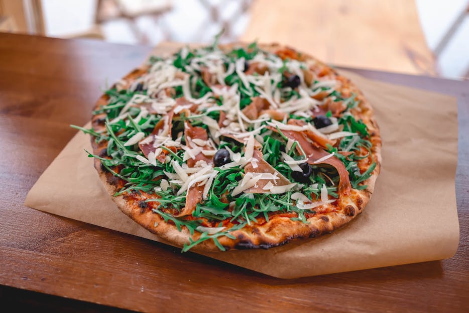 Pizzerija Plus Mouth-watering gourmet pizza topped with arugula, prosciutto, olives, and cheese on a wooden table.