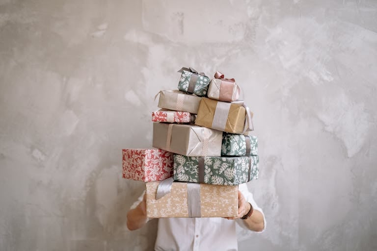 WhatUTalkingBoutWillis Gift Guide Person hiding behind a tall stack of gift-wrapped boxes against a gray wall, indoors.