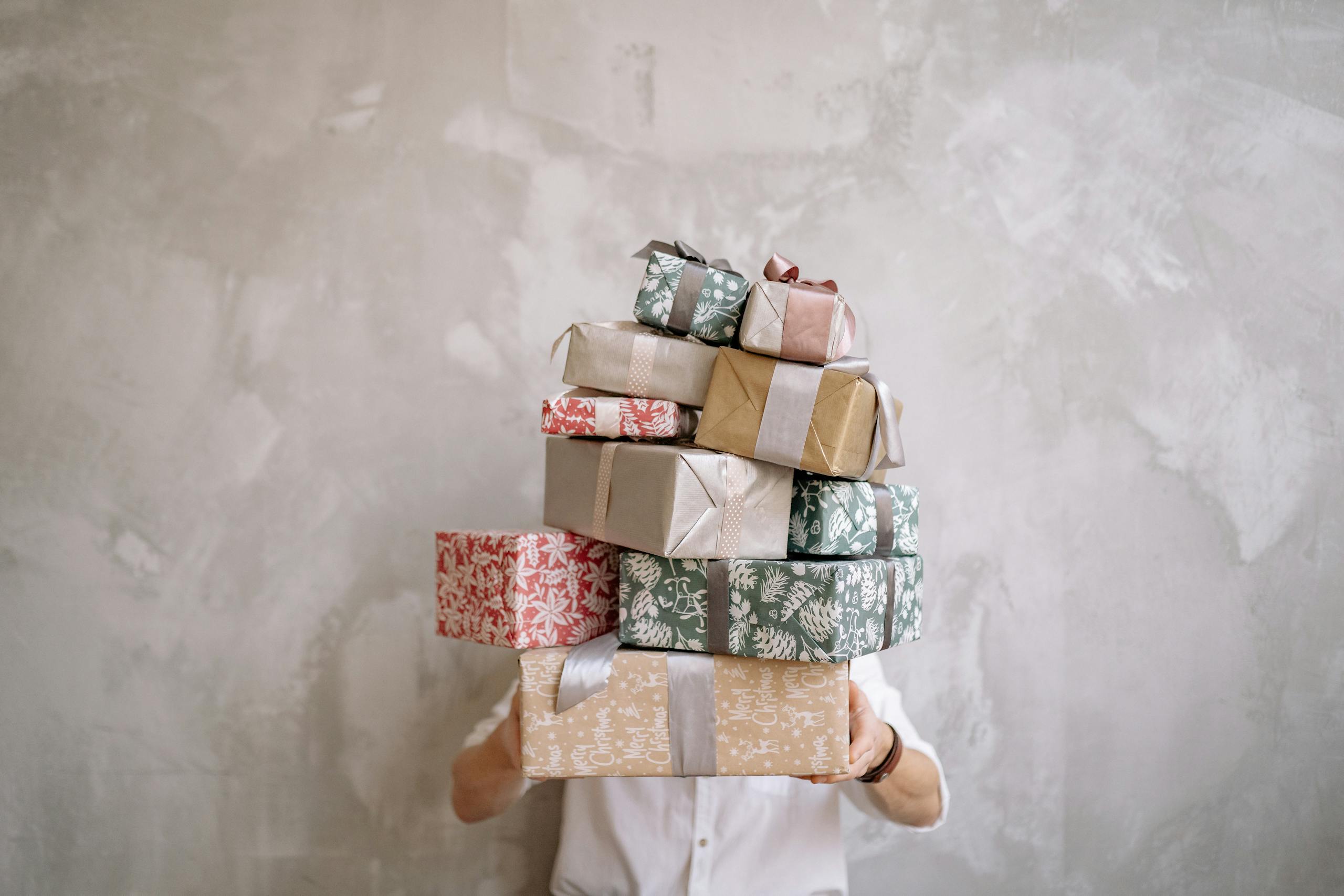 WhatUTalkingBoutWillis Gift Guide Person hiding behind a tall stack of gift-wrapped boxes against a gray wall, indoors.