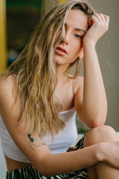 Laryssa Farmiga Portrait of a blonde woman with tattoos sitting and touching her forehead. Casual indoor setting.
