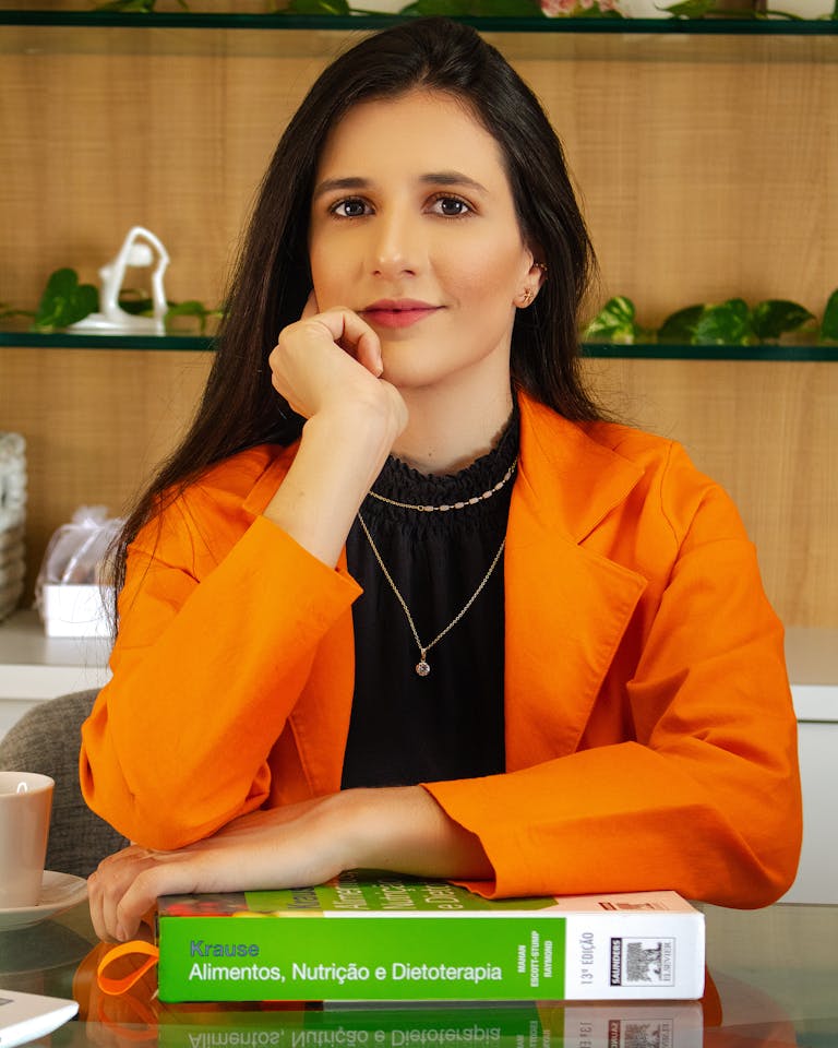 Laryssa Farmiga Woman in a vibrant orange jacket sits with a book on nutrition, looking confident.